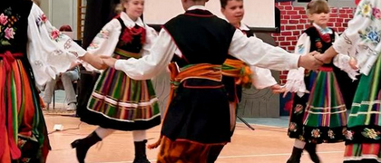 Na zdjęciu widać grupę młodych ludzi w tradycyjnych strojach ludowych, którzy wykonują taniec na sali gimnastycznej. Stroje są kolorowe, zdobione haftami, a dominują w nich barwy czerwieni, zieleni i czerni. Chłopcy mają na sobie białe koszule, czarne spodnie i haftowane kamizelki, a dziewczynki białe bluzki, haftowane gorsety i szerokie, wzorzyste spódnice. Tancerze trzymają się za ręce, tworząc okręgi lub inne figury taneczne, a ich pozy sugerują dynamiczny ruch. W tle widoczny jest parkiet sali gimnastycznej z namalowanymi liniami oraz elementy wyposażenia, takie jak tablica do koszykówki.