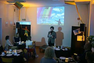 Zdjęcie przedstawiające osobę na scenie mówiącą do mikrofonu. W tle widok ekranu z projektora. Na pierwszym planie stoliki i siedzące osoby.