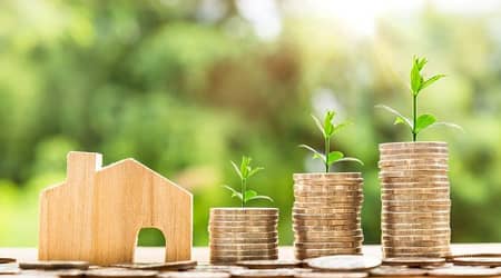 Monety ustawione w trzech stosach różnej wielkości. Obok drewniany domek.