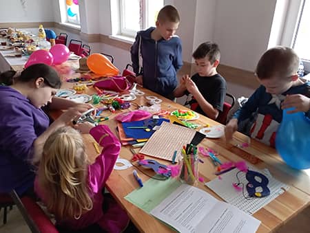  Zdjęcie przedstawiające dzieci tworzące maski karnawałowe przy stoliku.