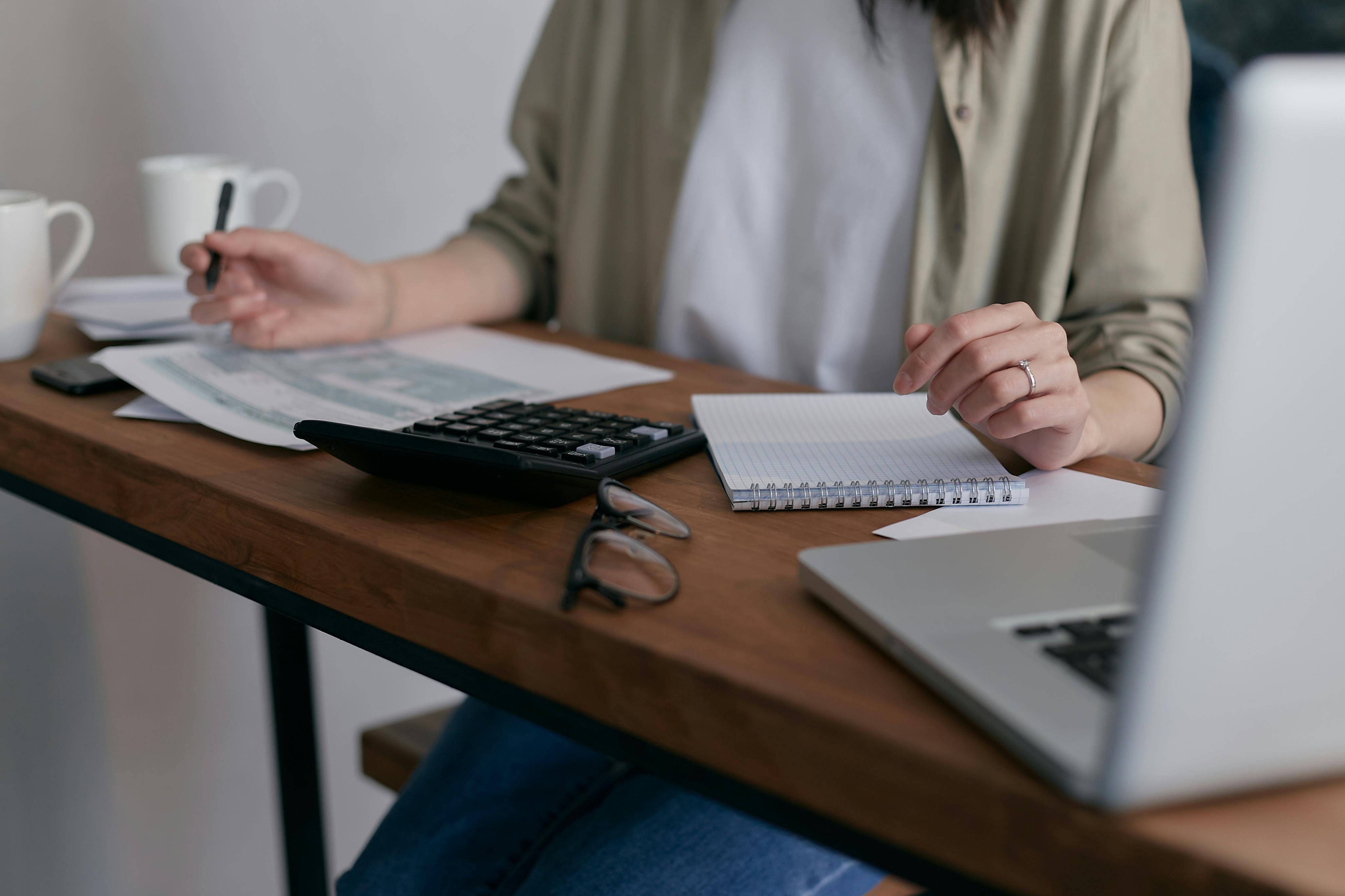 https://www.pexels.com obraz przedstawia panią przy stole z otwartym komputerem trzymającą długopis i liczącą