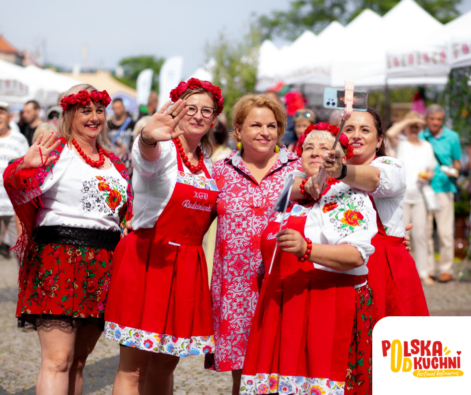 Na tym radosnym zdjęciu widzimy grupę pięciu kobiet, prawdopodobnie przyjaciółek lub uczestniczek jakiegoś wydarzenia kulturalnego. Cztery z nich ubrane są w podobne stroje, składające się z białych bluzek z czerwonymi haftowanymi wzorami kwiatowymi, czerwonych spódnic z kolorowymi paskami na dole oraz czerwonych wianków na głowach. Dwie z nich mają na sobie czerwone fartuchy z białym napisem "KGW Rudzienice". Jedna z kobiet w pierwszym rzędzie trzyma w ręku polską flagę i uśmiecha się do kamery, robiąc sobie selfie telefonem przymocowanym do kijka. Pozostałe kobiety również pozują, machając do obiektywu.  Piąta kobieta, stojąca w środku, ubrana jest w wzorzystą sukienkę w odcieniach czerwieni i bieli. Wszystkie kobiety wyglądają na szczęśliwe i dobrze się bawiące.  W tle widać rozmyty tłum ludzi, stoiska i białe namioty, co sugeruje, że zdjęcie zostało zrobione podczas jakiegoś festynu, jarmarku lub innego wydarzenia plenerowego. W prawym dolnym rogu widoczne jest logo "POLSKA OD KUCHNI". Cała scena jest pełna pozytywnej energii i patriotycznego ducha.