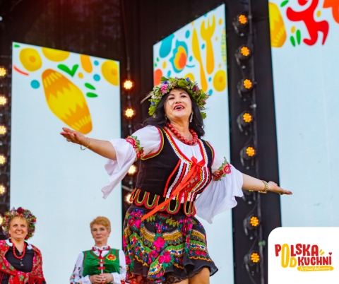 źródło: Festiwal „Polska od Kuchni” Na pierwszym planie widzimy uśmiechniętą kobietę w kwiecistym wieńcu na głowie, tradycyjnym polskim stroju ludowym, tańczącą z rozłożonymi ramionami. Jej czarna kamizelka jest bogato zdobiona kolorowymi haftami i czerwonymi wstążkami. Spódnica, również wzorzysta w kwiaty, dodaje dynamiki jej ruchom. Na szyi ma kilka sznurów czerwonych korali. W tle, po lewej stronie, widoczne są dwie inne kobiety, również ubrane w regionalne stroje i wieńce, wyglądające na to, że śpiewają lub obserwują taniec. Za nimi rozciąga się jasne tło, prawdopodobnie scena, z dekoracyjnymi elementami graficznymi w górnej części. Po prawej stronie kadru znajduje się logo "POLSKA OD KUCHNI". Całość zdjęcia emanuje radością i energią, sugerując występ folklorystyczny lub festiwal kultury ludowej. Jasne światło i żywe kolory podkreślają świąteczny charakter sceny.